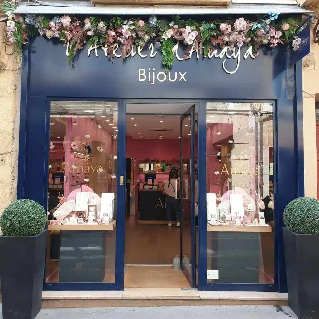 Façade bleue de L'Atelier d'Amaya Bijoux, décorée d'une guirlande florale. Une vitrine exposant des bijoux et deux plantes vertes encadrent l'entrée.