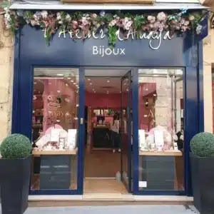 Façade bleue de L'Atelier d'Amaya Bijoux, décorée d'une guirlande florale. Une vitrine exposant des bijoux et deux plantes vertes encadrent l'entrée.