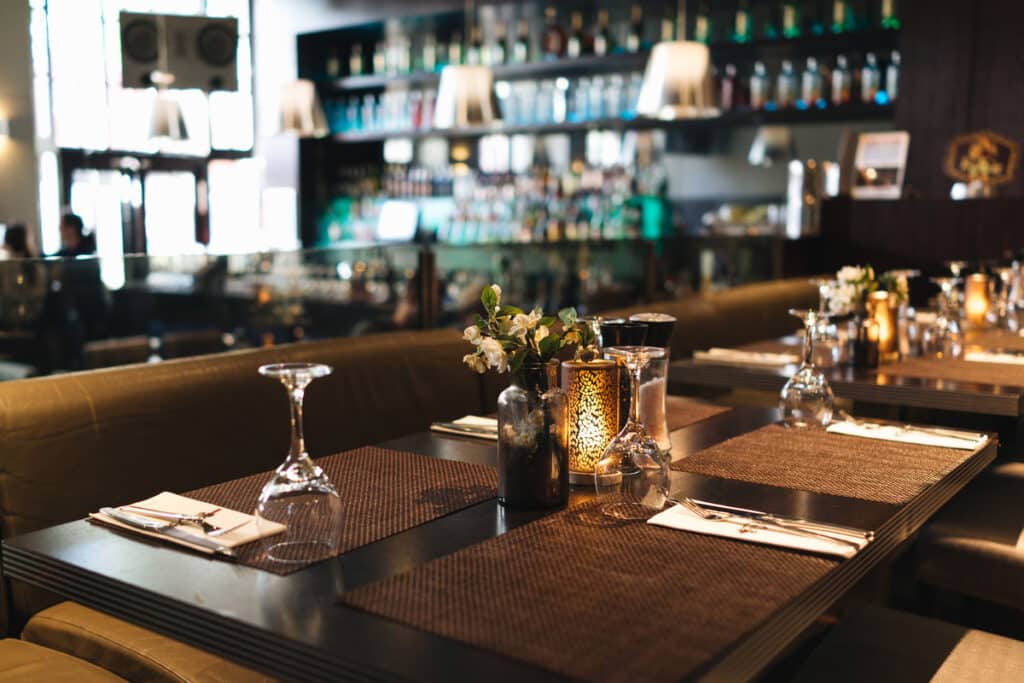 Table de restaurant dressée avec verres, fleurs et bougie. Bar moderne éclairé en arrière-plan. Ambiance chaleureuse et chic.